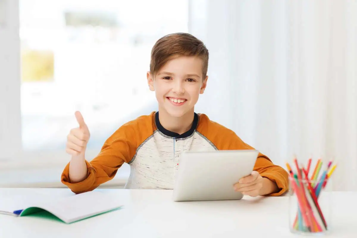 Smiling child giving a thumbs-up while holding a tablet, sitting at a desk with a notebook and colored pencils, illustrating the positive impact of homeschooling on student happiness and engagement.