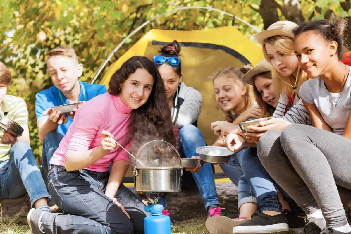 A group of children camping, cooking together in front of a tent, highlighting outdoor learning and teamwork for homeschool socialization