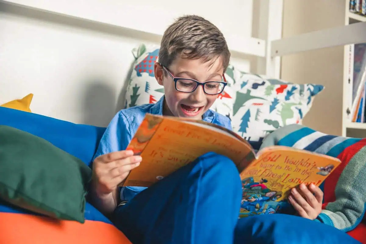 Young boy with glasses sitting on a colorful couch, smiling as he reads a book titled 'You Wouldn't Want to Be in the Great Fire of London!'. The background shows patterned pillows with trees and a bookshelf.