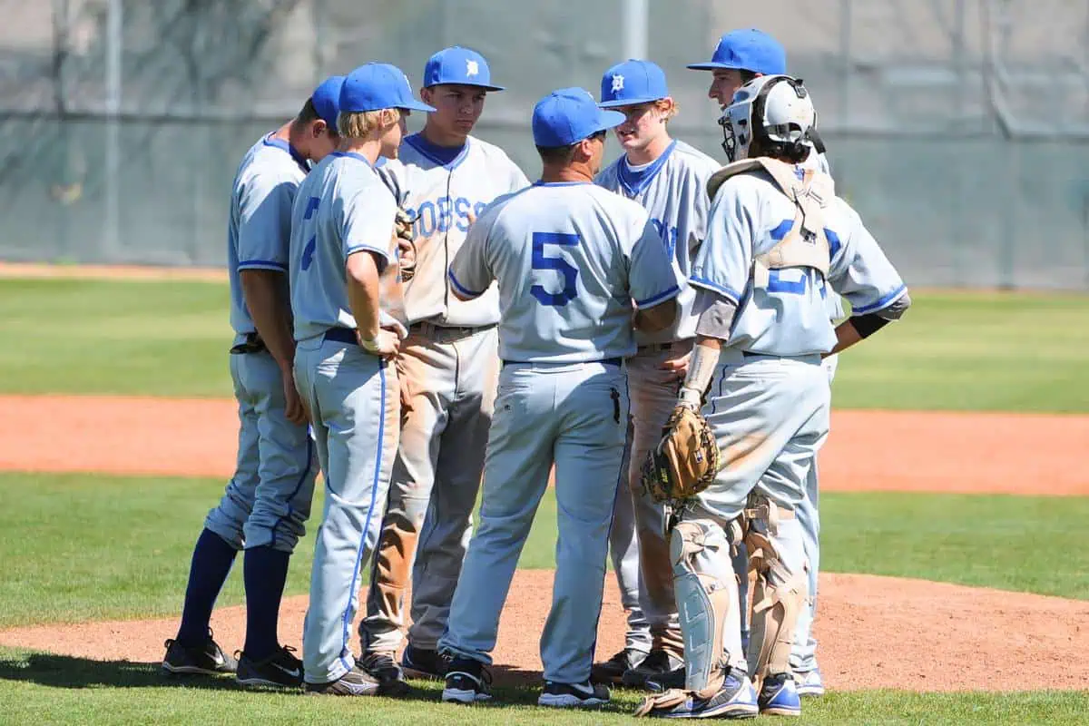 A high school baseball team, dressed in light blue and white uniforms, gathers on the pitcher's mound for a discussion during a game. The keyword for this image is homeschool pros and cons, which may be discussed in the context of extracurricular activities like sports.