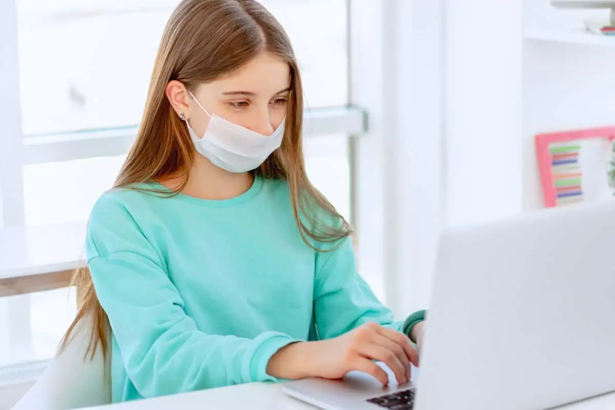 A young girl wearing a mask and working on a laptop, highlighting the concept of continuing education from home despite being sick.