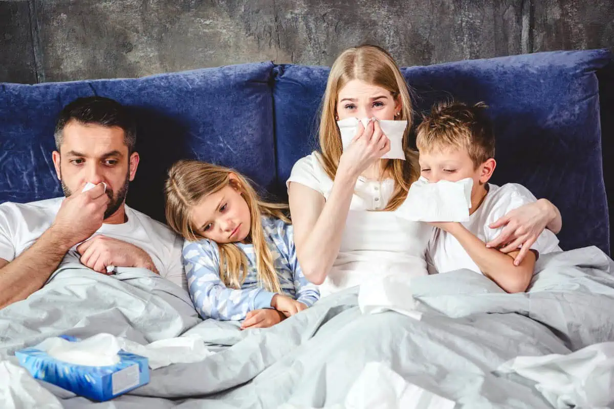 A family of four, all appearing sick, sitting in bed with tissues. The father, mother, and two children look tired and are using tissues to blow their noses.