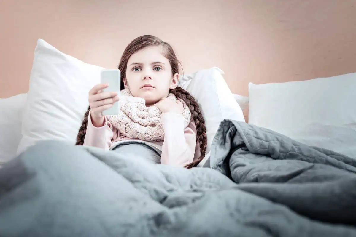 A young girl lying in bed, holding a remote control, wrapped in a scarf, and looking unwell, suggesting she is resting and watching TV during a sick day.