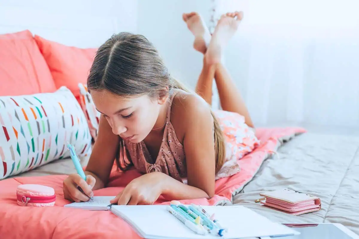 fun gratitude journal prompts for kids with a girl sitting on a bed writing in her gratitude journal notebook.