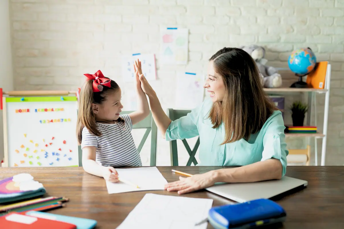 A smiling woman and a young girl with a red bow in her hair give each other a high-five over a homeschooling setup, featuring workbooks and a laptop. In the background, a whiteboard reads 'Home Schooling' with educational posters and a globe, illustrating a supportive home education environment. Perfect imagery for discussing the 'best free homeschool curriculum'.