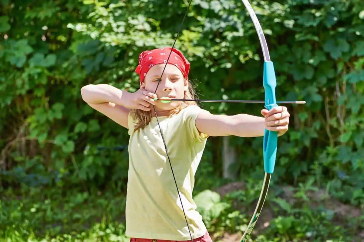 Teenager aiming a bow and arrow outdoors, wearing a red bandana, showcasing an exciting and adventurous activity