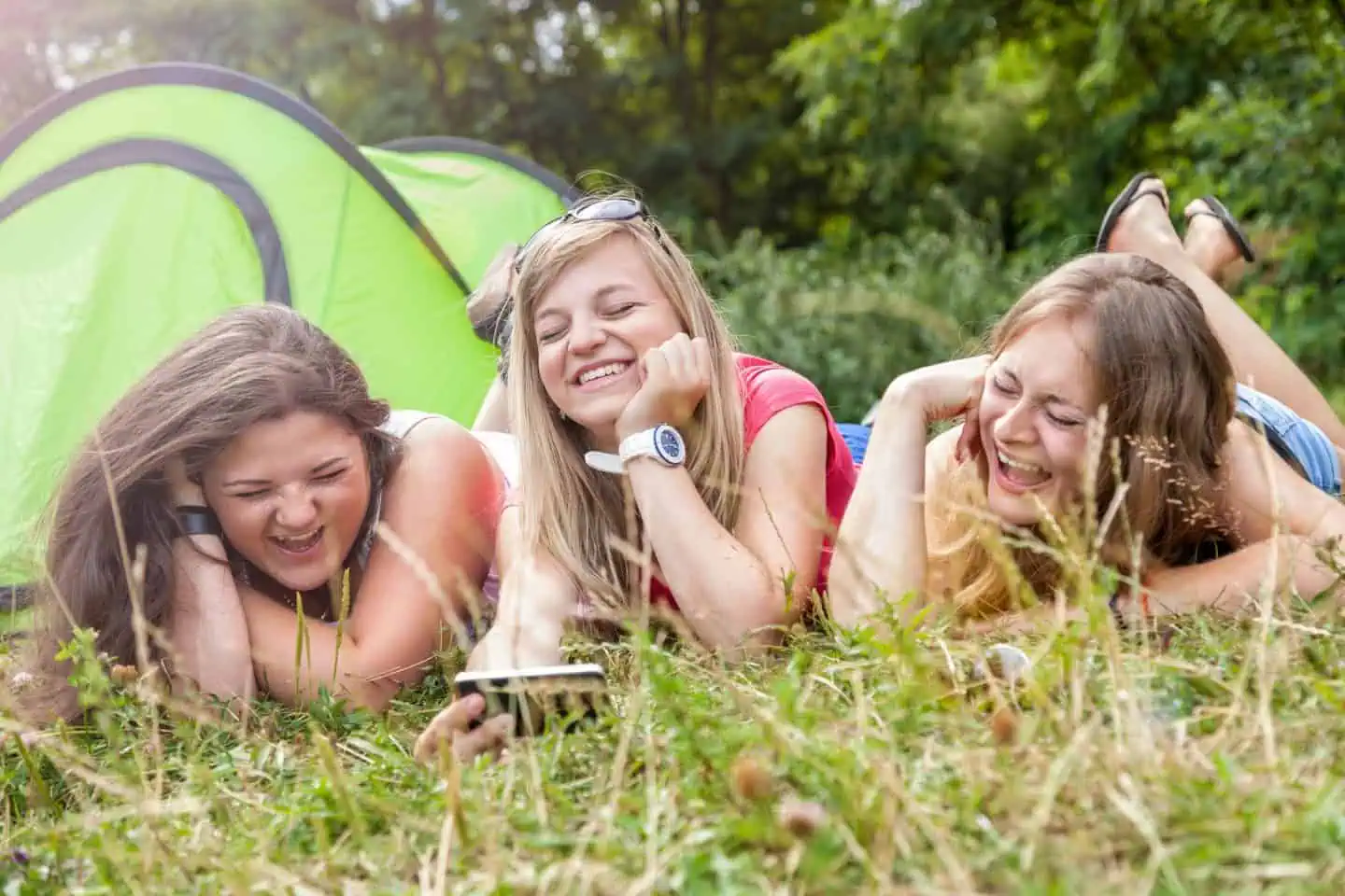 Three teenage girls lying on the grass in front of a green tent, laughing and looking at a smartphone.