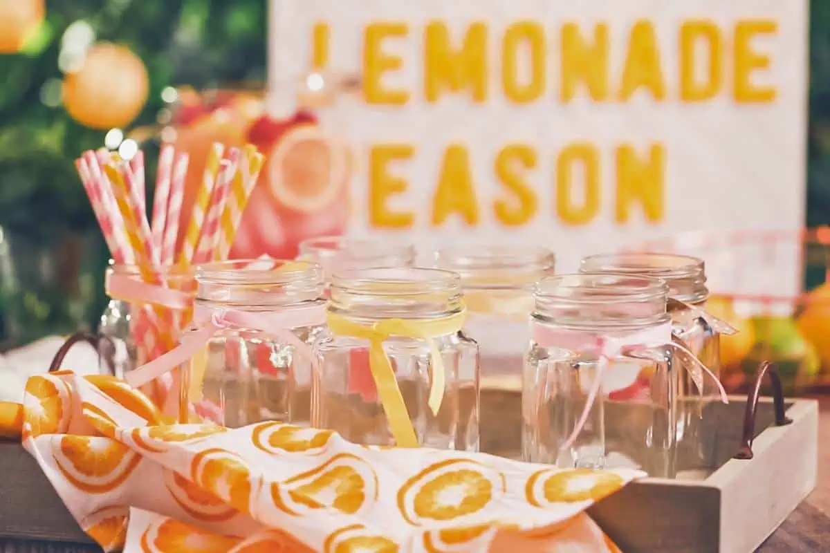 Mason jars with pastel-colored ribbons and striped straws arranged on a table with a sign in the background that reads 'Lemonade Season,' perfect for a summer fun.