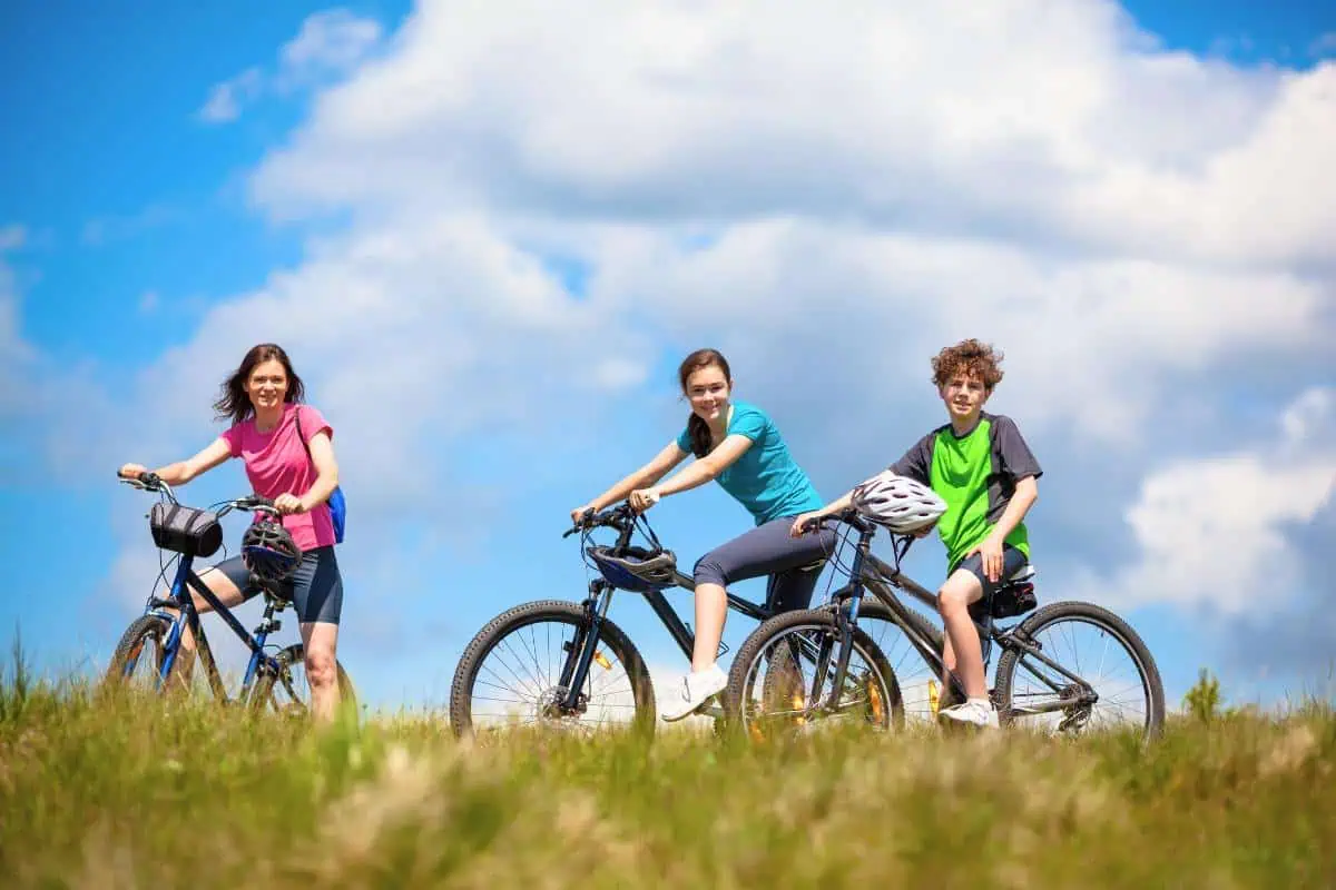 Three teenagers on mountain bikes stopping on a grassy hill under a blue sky with clouds, highlighting an adventurous activity.