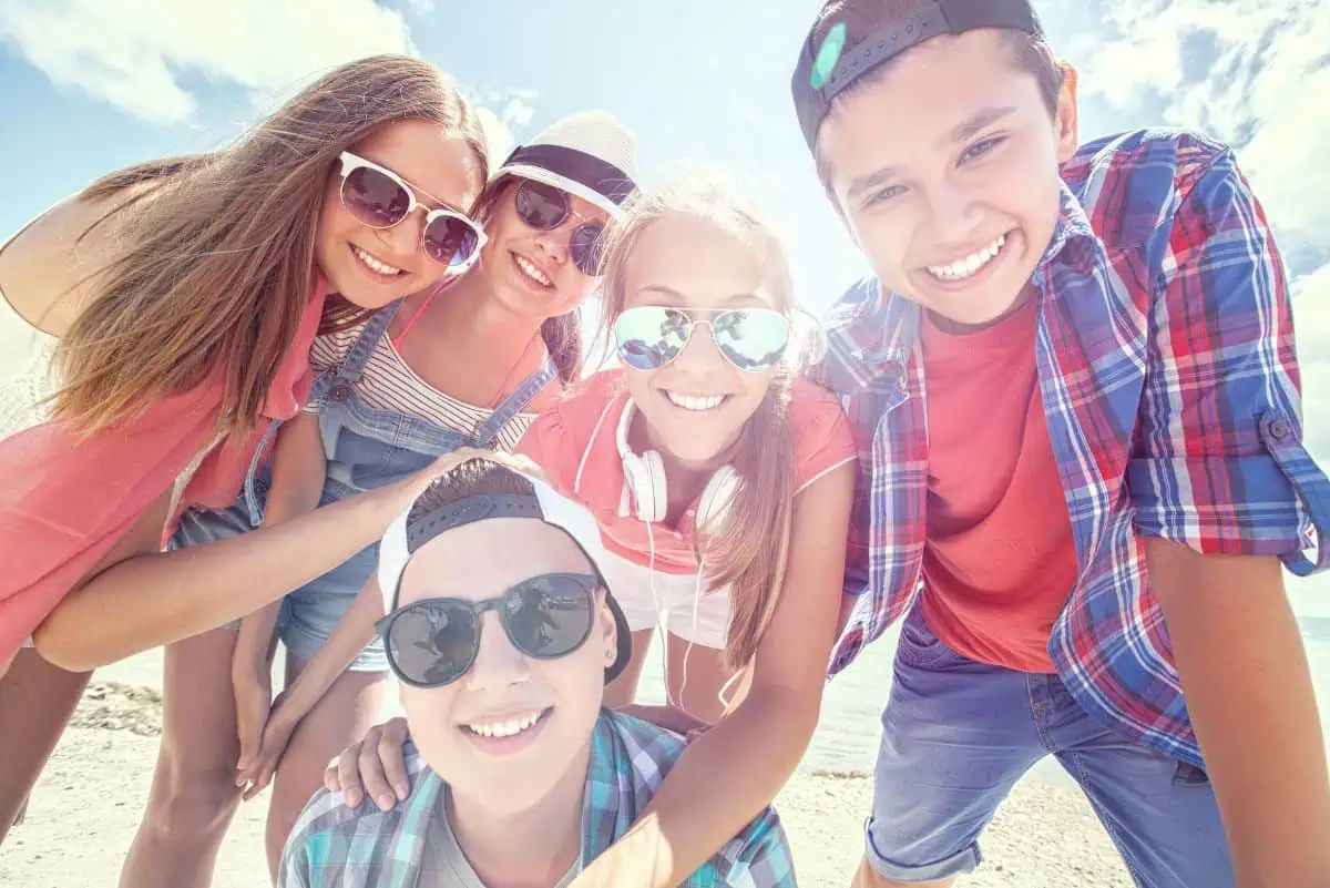 Five teenagers smiling and posing together at the beach, all wearing sunglasses and enjoying the sunny day.