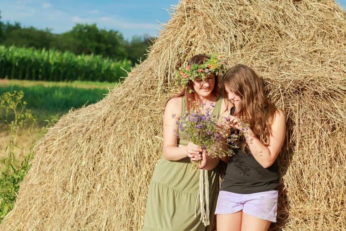 Two teenage girls standing by a haystack in a field, one wearing a flower crown and holding a bouquet, enjoying a summer day. Ideal for a summer bucket list for teenagers.