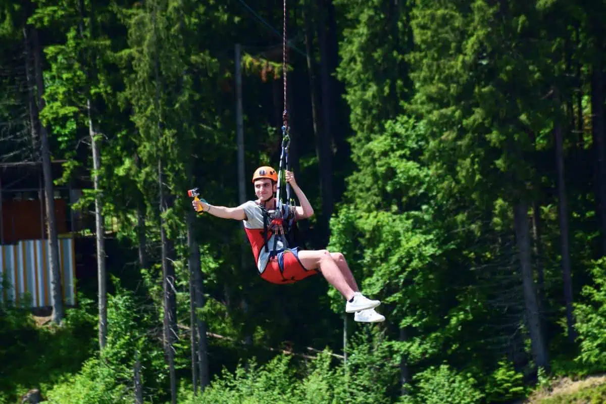 Teenager zip-lining through a forest, holding a camera and smiling, showcasing an adventurous activity