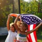 Two children joyfully running with an American flag, celebrating the 4th of July. The image promotes unforgettable 4th of July games for kids, perfect for family fun and outdoor activities. The bright summer day and festive spirit capture the essence of Independence Day celebrations