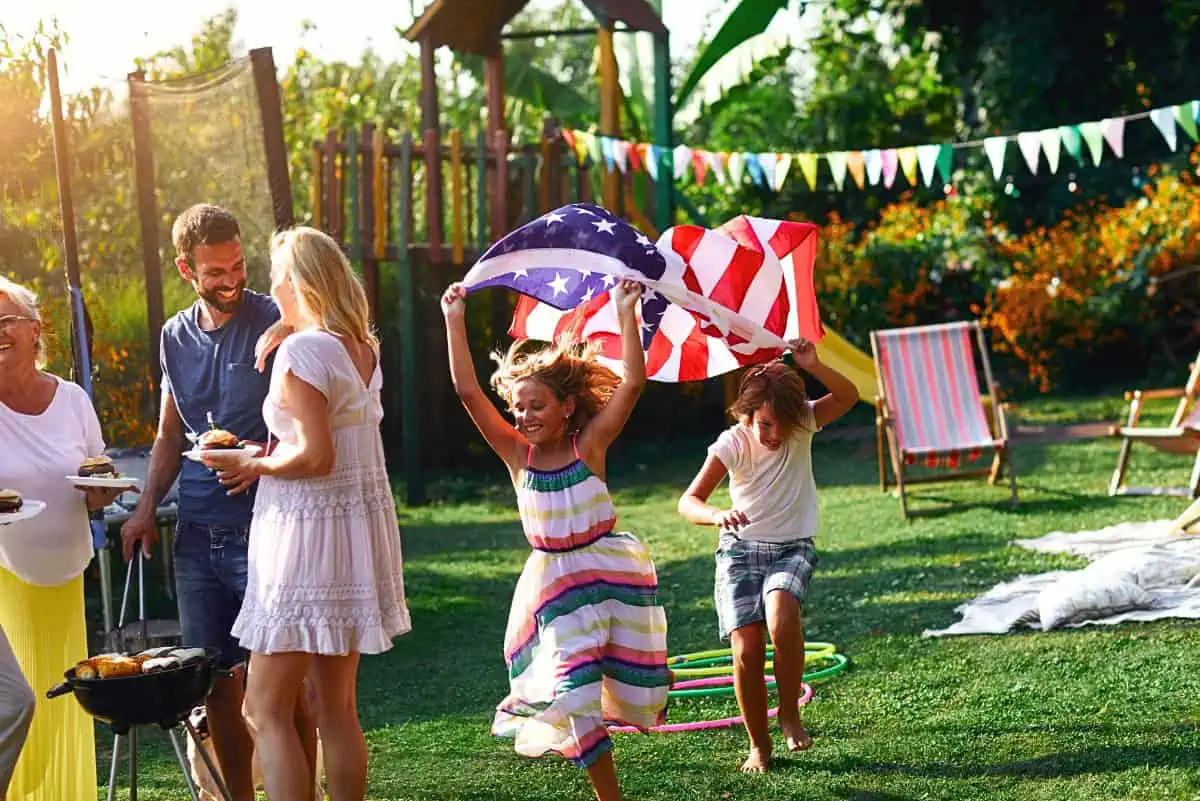 A lively 4th of July celebration with family and friends enjoying outdoor activities. Children run and play with an American flag, while adults gather around a grill with food and smiles. Bright summer decorations and a sunny day set the perfect scene for festive 4th of July games.