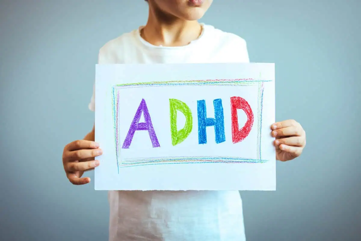 A child holding a white sign with "ADHD" written in large, colorful letters against a plain background. The letters "A," "D," "H," and "D" are in red, green, blue, and purple, respectively.