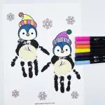 penguin handprint art with 2 painted handprint penguins wearing colorful hats