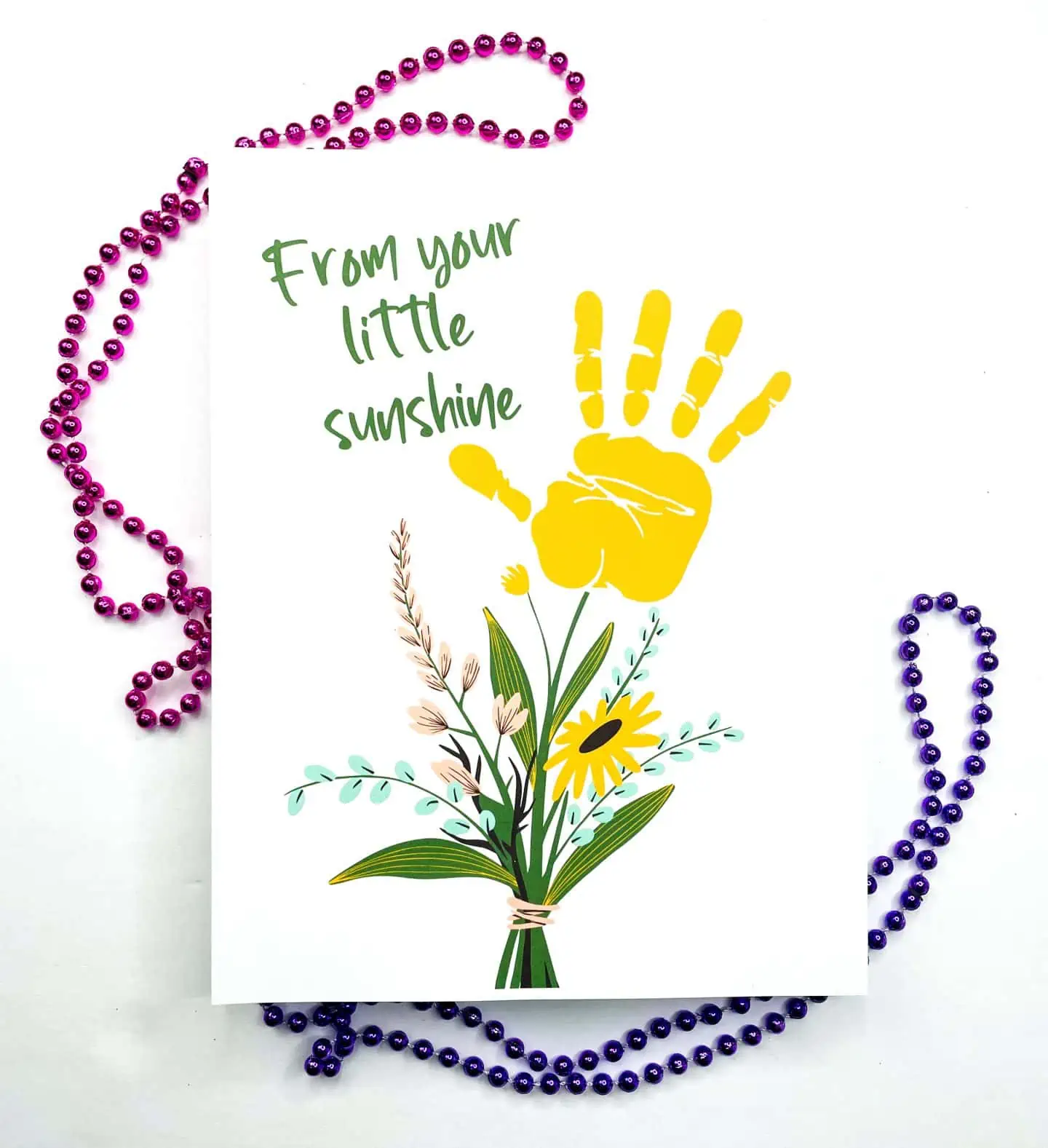 The image displays a handprint art printable that reads "From your little sunshine" in green lettering, featuring a bright yellow handprint above a colorful illustration of a floral bouquet. The artwork is embellished by a framing of purple and blue bead necklaces on a white background.


