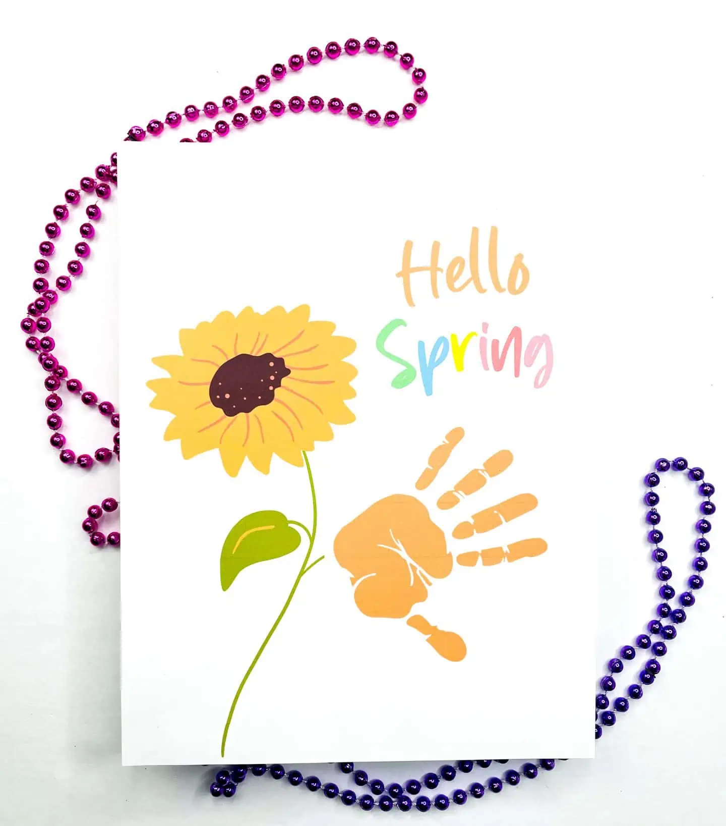 A festive "Hello Spring" handprint art printable with a large yellow sunflower and a child's orange handprint, flanked by purple bead necklaces.

