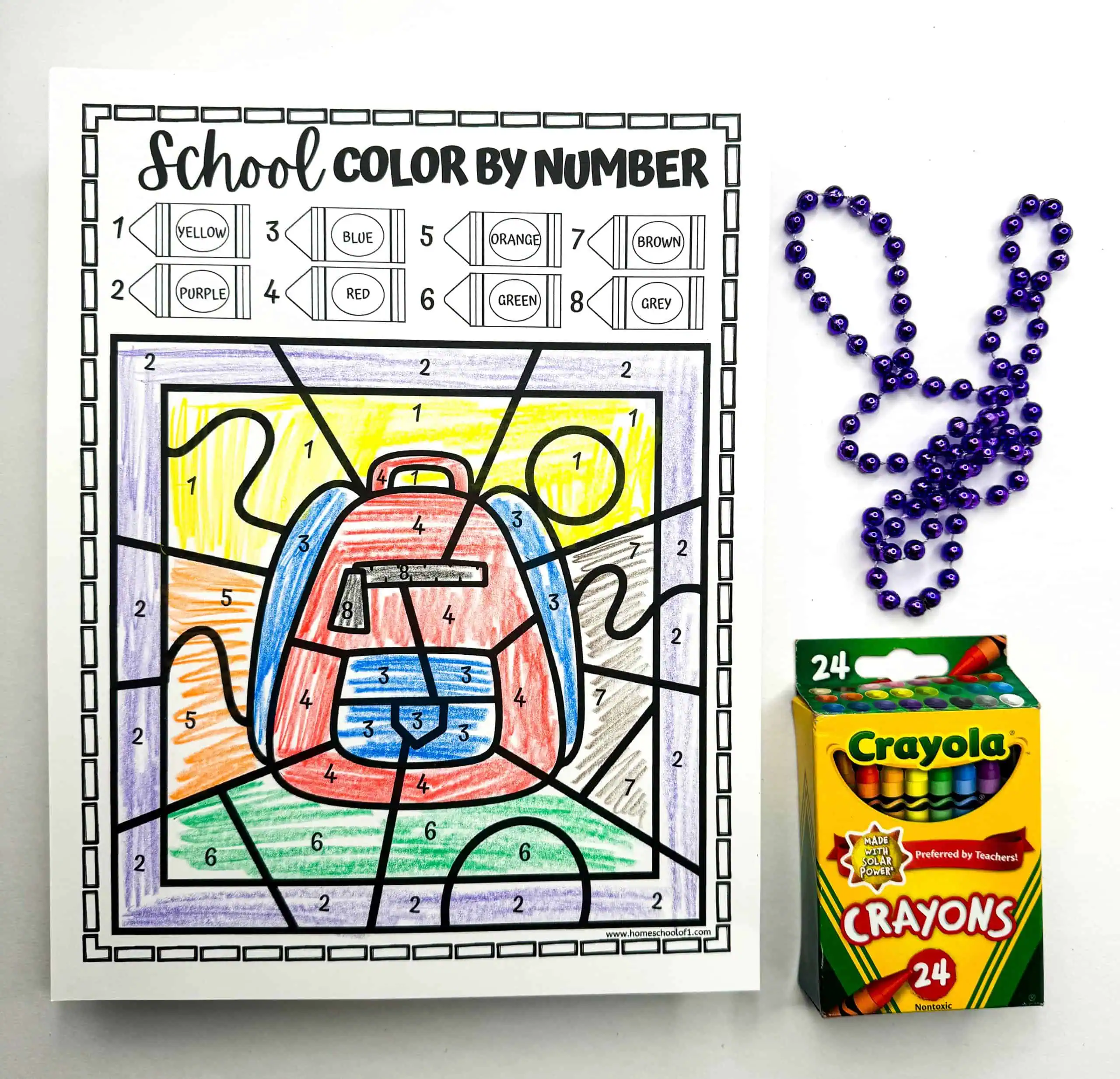 A color by number sheet presents a vibrantly colored school backpack. Alongside the completed worksheet are purple beads arranged as a rabbit and a box of Crayola crayons, highlighting this as a playful learning activity.