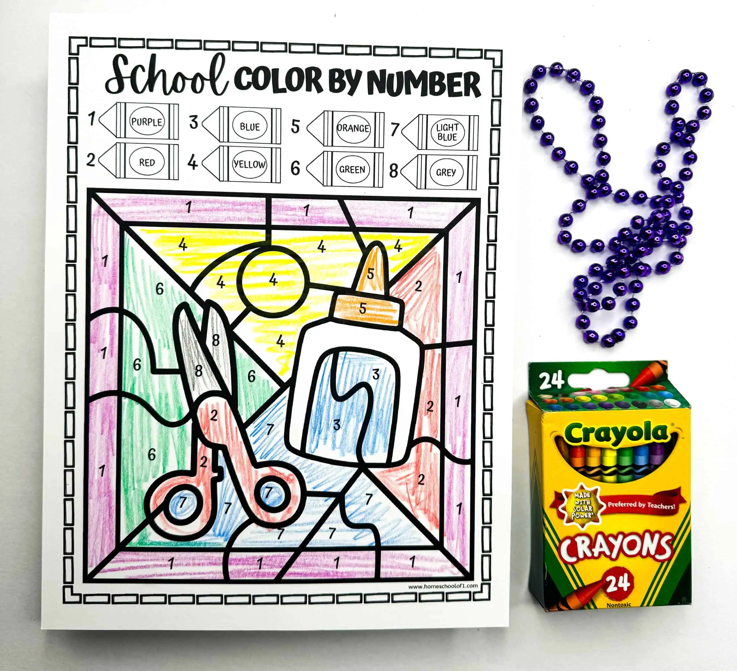 A color by number worksheet features various school stationery items, each shaded according to a color key. Decorative beads in a rabbit shape and a box of Crayola crayons complete this educational coloring scene.