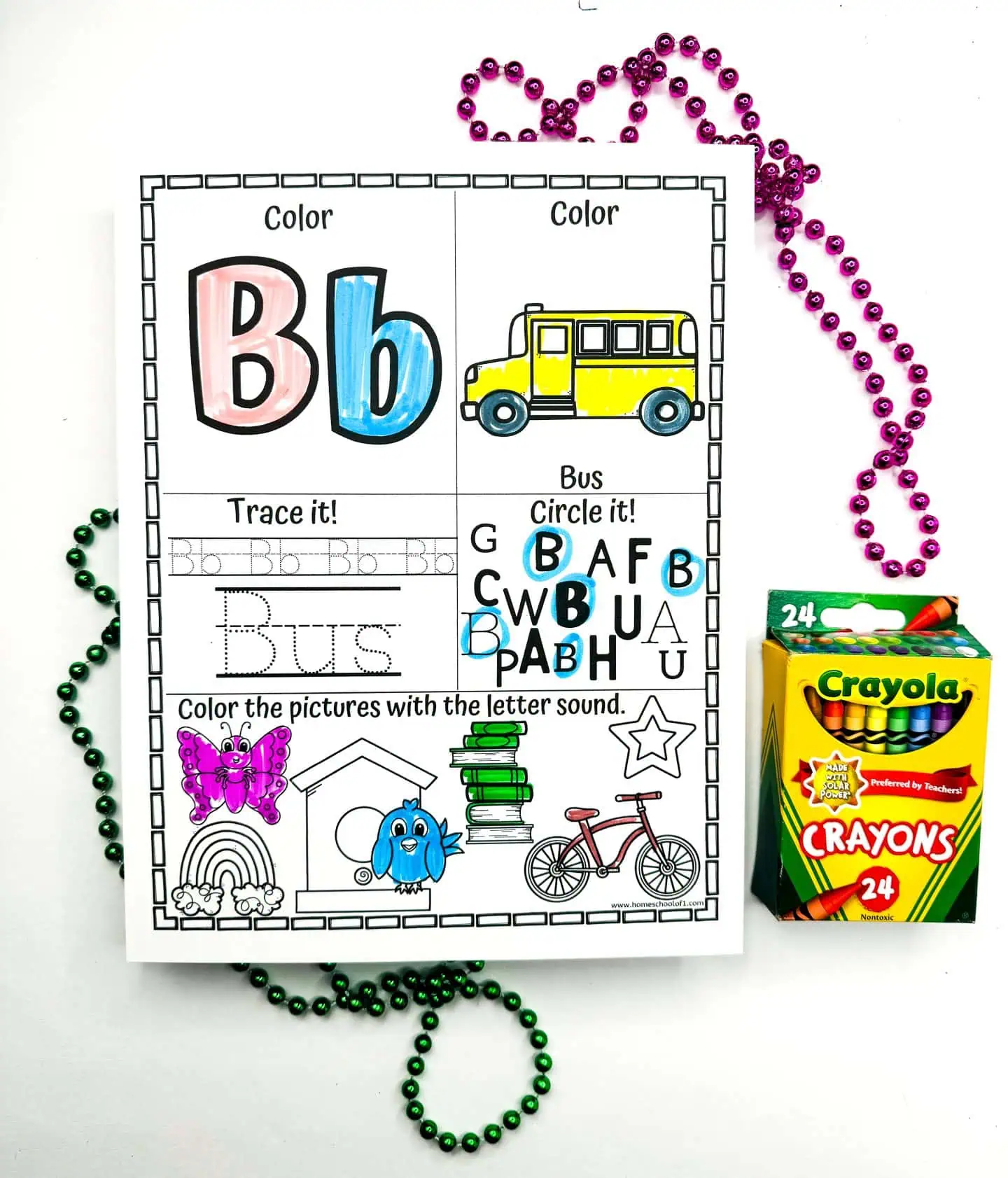 An educational worksheet featuring the letter B with sections for coloring and tracing the word "Bus." The worksheet is partially colored and depicts a yellow school bus, along with various items starting with B, like a butterfly and books. Green and purple beads are artistically arranged around it, with a box of Crayola crayons to the side.