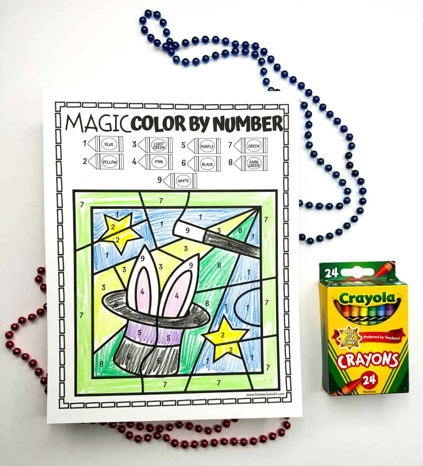 Color by number activity sheet featuring a drawing of a magic hat with bunny ears popping out, surrounded by colorful beads and a box of Crayola crayons on a white surface.