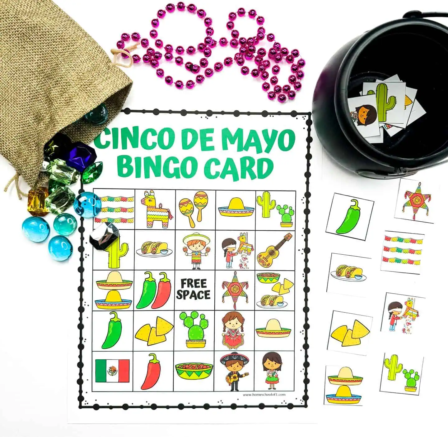 Colorful 'Cinco de Mayo Bingo Card' displayed with purple beads forming the word 'Olay,' alongside a burlap sack with assorted game gems and a black cauldron of bingo cards, set on a white surface.