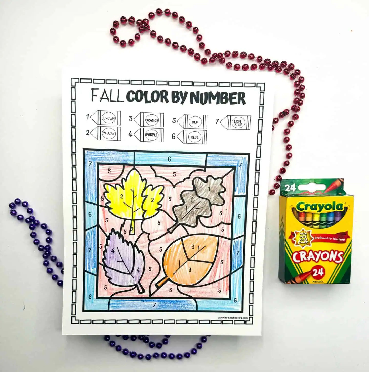 Worksheet showcasing leaves in a color by number activity for fall, accented with a Crayola crayon box and decorative bead necklaces.