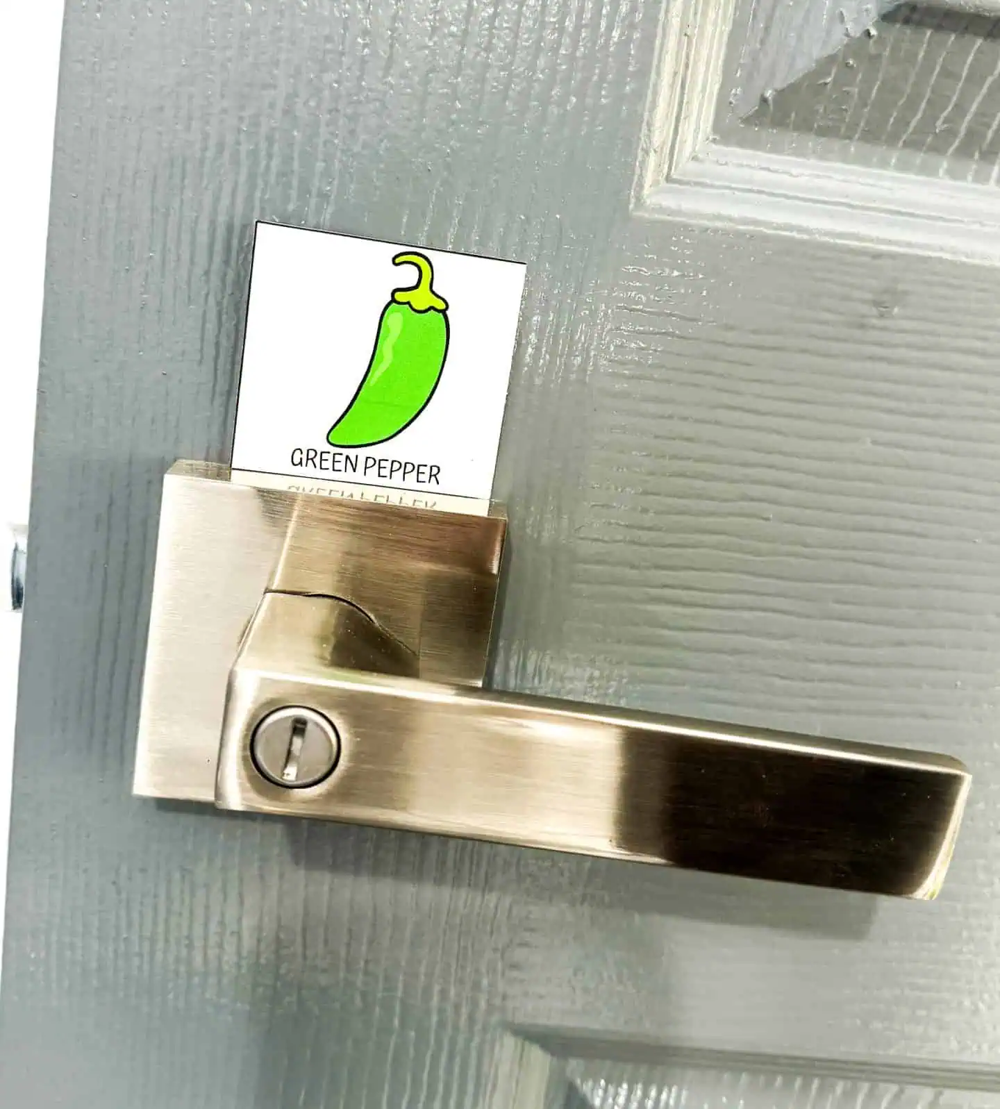 A modern, satin nickel door handle with a 'Cinco de Mayo Scavenger Hunt' game card of a green pepper slipped into the handle’s lock, set against a textured gray door.