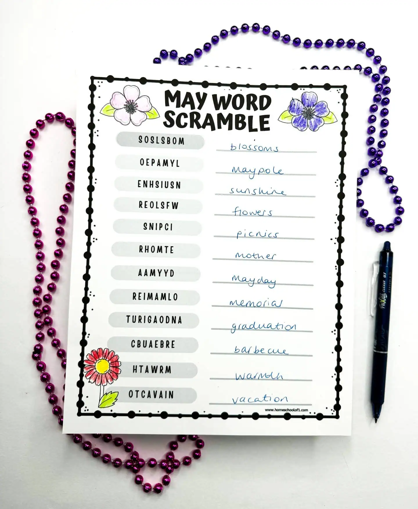 Challenging May Word Scramble sheet with scrambled words like 'blossoms' and 'vacation' alongside their solved counterparts, surrounded by Mardi Gras beads and a blue pen