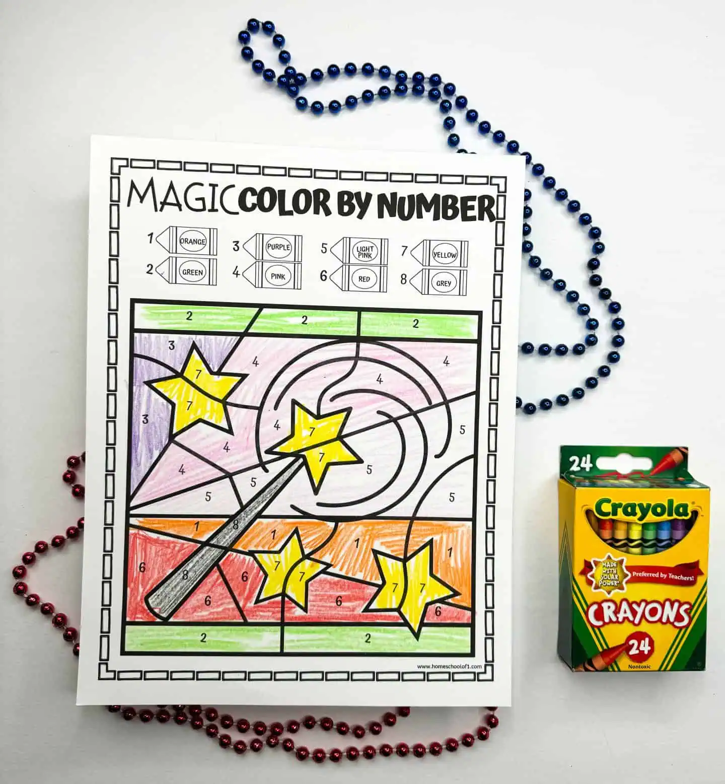 A child's completed color by number worksheet showcasing a magic wand over a swirling background, next to a string of blue beads and a Crayola crayon box.