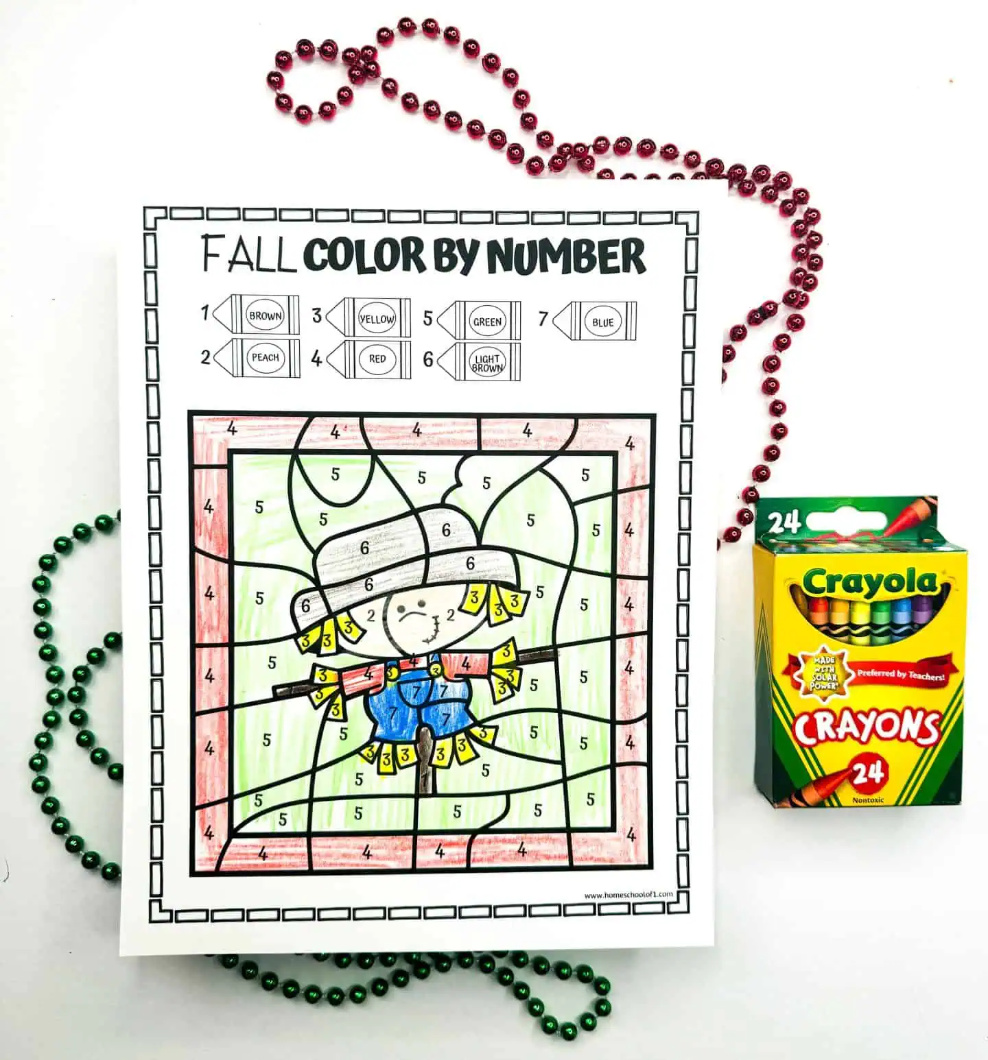 Color by number page displaying a friendly scarecrow, complemented by green bead necklaces and a 24-pack of Crayola crayons.