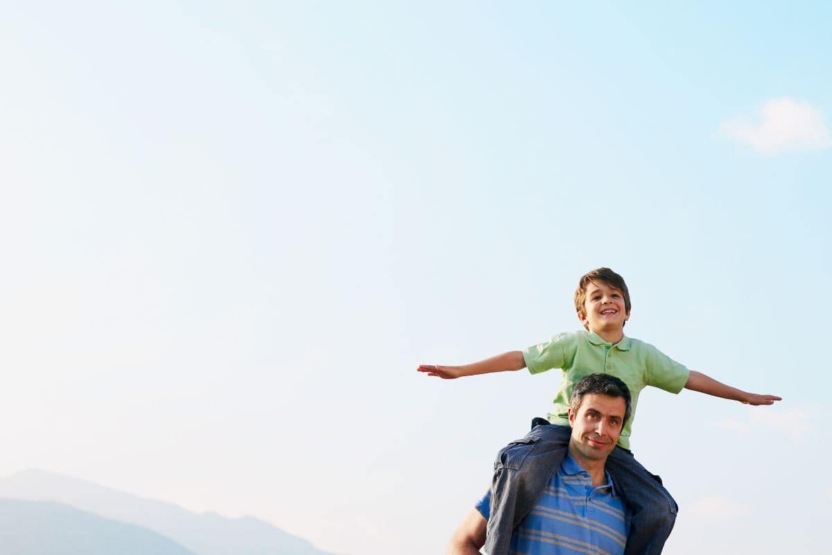 A happy child with arms outstretched sits on a man's shoulders, both smiling against a clear blue sky backdrop.