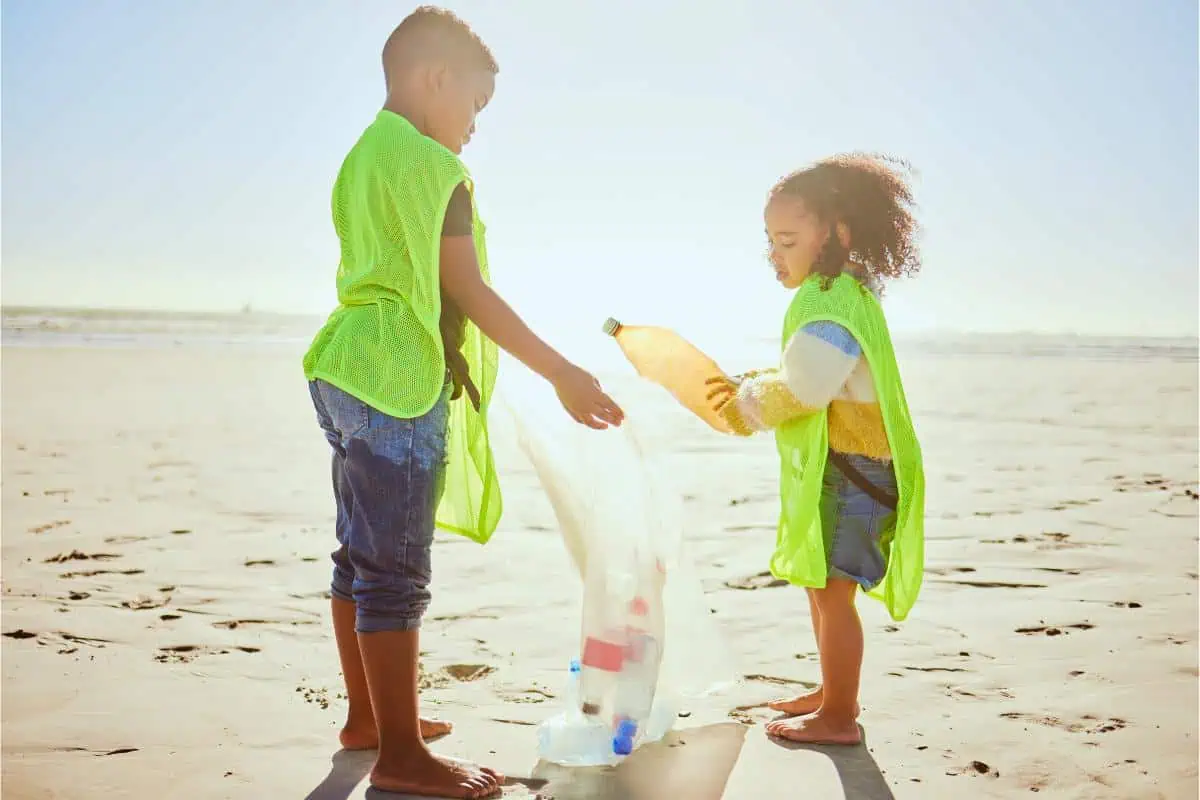 Two children wearing neon vests pick up plastic bottles on a beach, highlighting environmental stewardship and summer learning through community service.