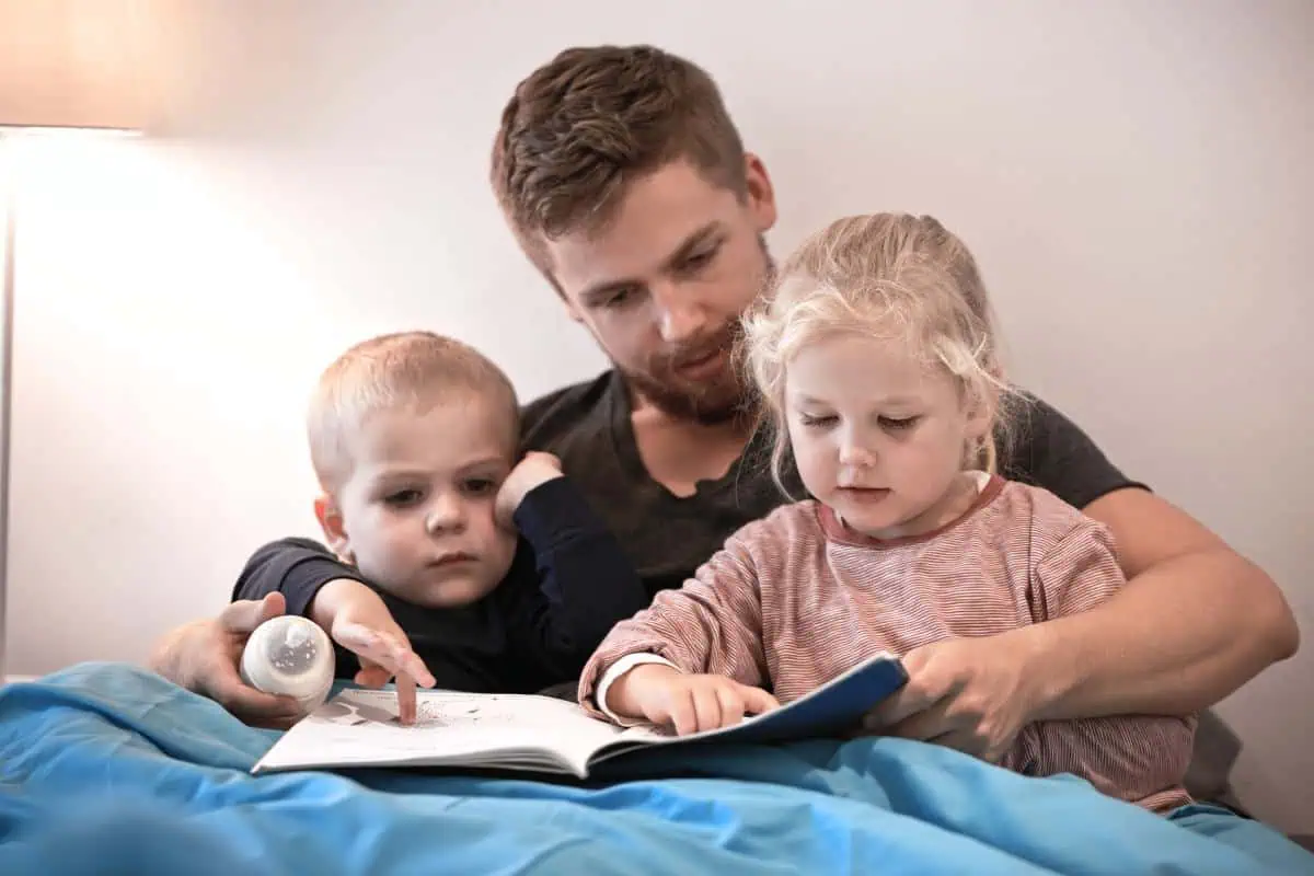 A father is reading a book with his two young children, both engaged and pointing at the pages. A wonderful moment to celebrate with Father's Day books for kids, highlighting family bonding and shared stories.