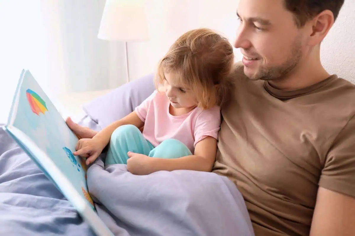 A dad and his daughter are reading a colorful picture book while snuggled up in bed. Ideal for bedtime stories on Father's Day.