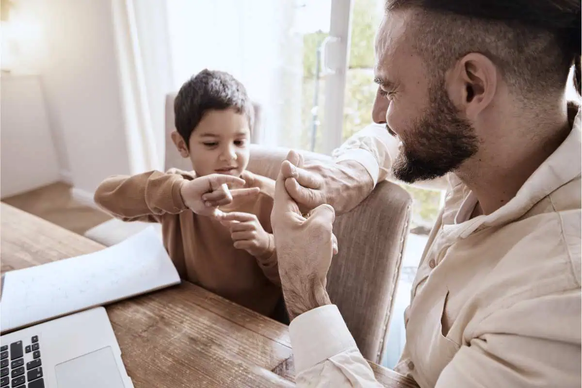 This image depicts a young boy and an adult man, possibly his father, engaging in an interactive activity. They are seated at a wooden table with a laptop open and a book visible, suggesting they might be learning or teaching something, potentially sign language. The boy is focused and pointing to his hand while the man smiles, guiding the boy's hand with a gentle touch. The room is well-lit with natural light streaming in from a window in the background, creating a warm and engaging learning environment.






