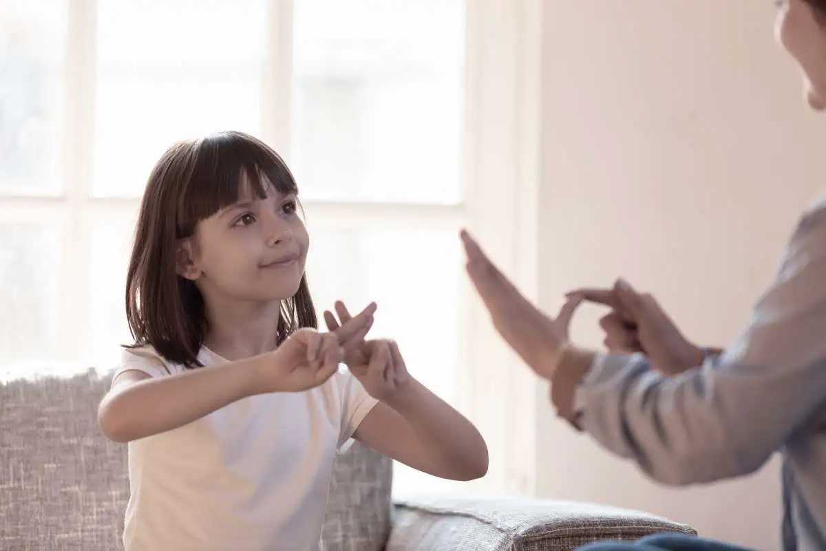 This image captures a tender moment between a young girl with shoulder-length hair and an adult, likely engaging in a sign language lesson. The child, dressed in a simple white shirt, focuses intently on forming hand signs, mirroring the gestures of the adult whose hands are visible in the frame. They are seated in a warmly lit room with soft natural light filtering through a window, emphasizing the serene and focused atmosphere of their learning experience.