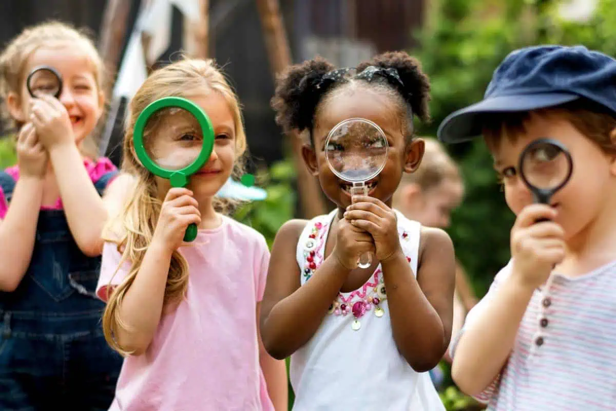 Group of children smiling and holding magnifying glasses, emphasizing hands-on exploration and summer learning through nature activities.