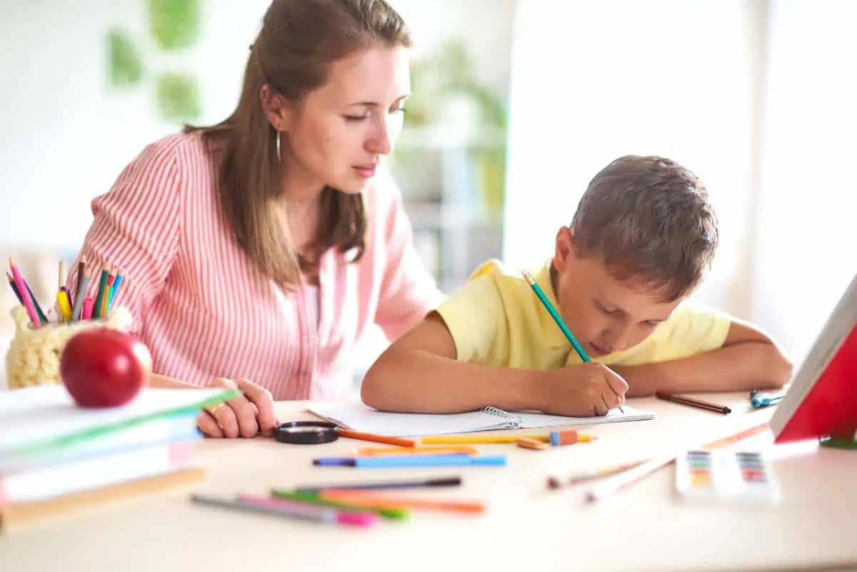 Mother homeschooling her child, sitting at a table with various school supplies, as the child concentrates on writing in a notebook, showcasing a dedicated and personalized learning environment.